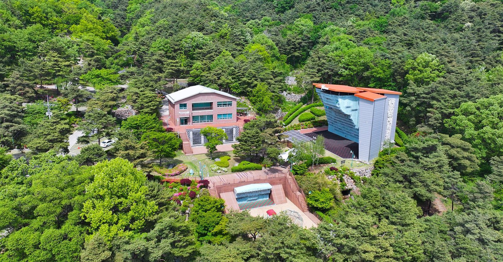 숲에 둘러싸인 붉은 벽돌 2층 건물과 주황 지붕을 얹은 높은 인공등반벽, 그 아래 작은 실외 등반벽을 위에서 내려다본 모습