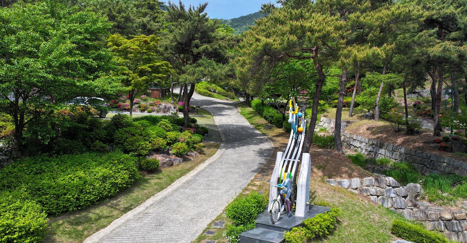소나무 사이로 굽은 포장 진입로와 자전거 조형물, 철쭉 화단